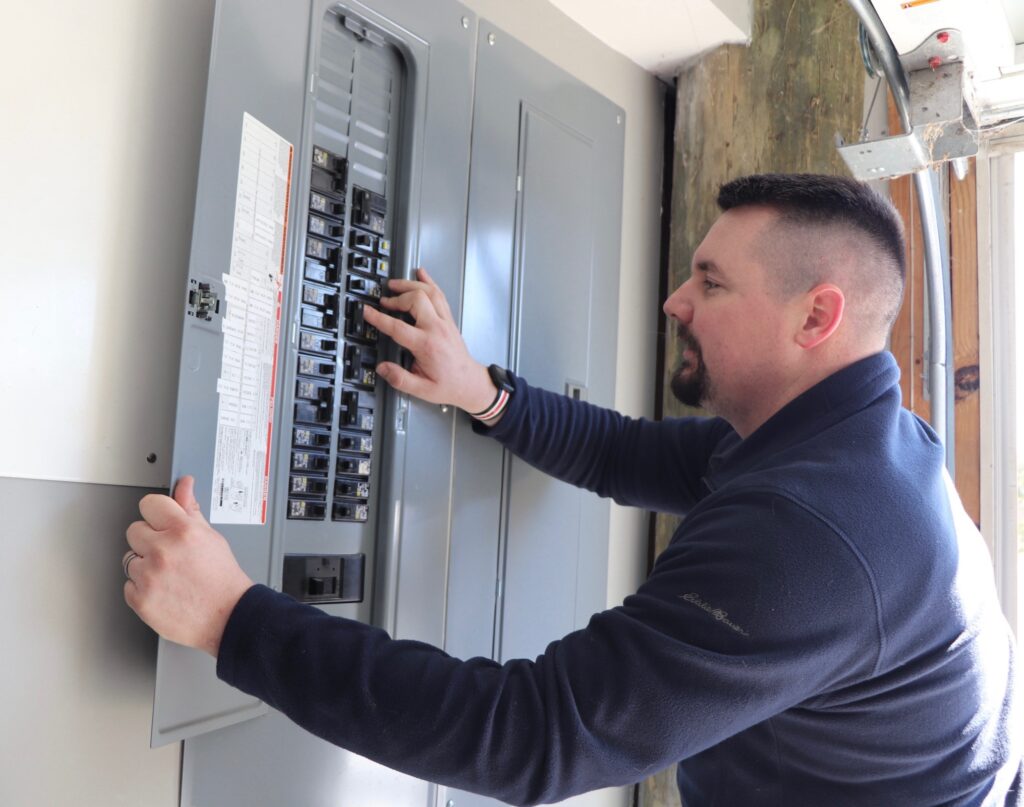 A man in a navy sweater adjusts switches inside a gray electrical breaker panel mounted on a wall, with sunlight coming in from the right side.