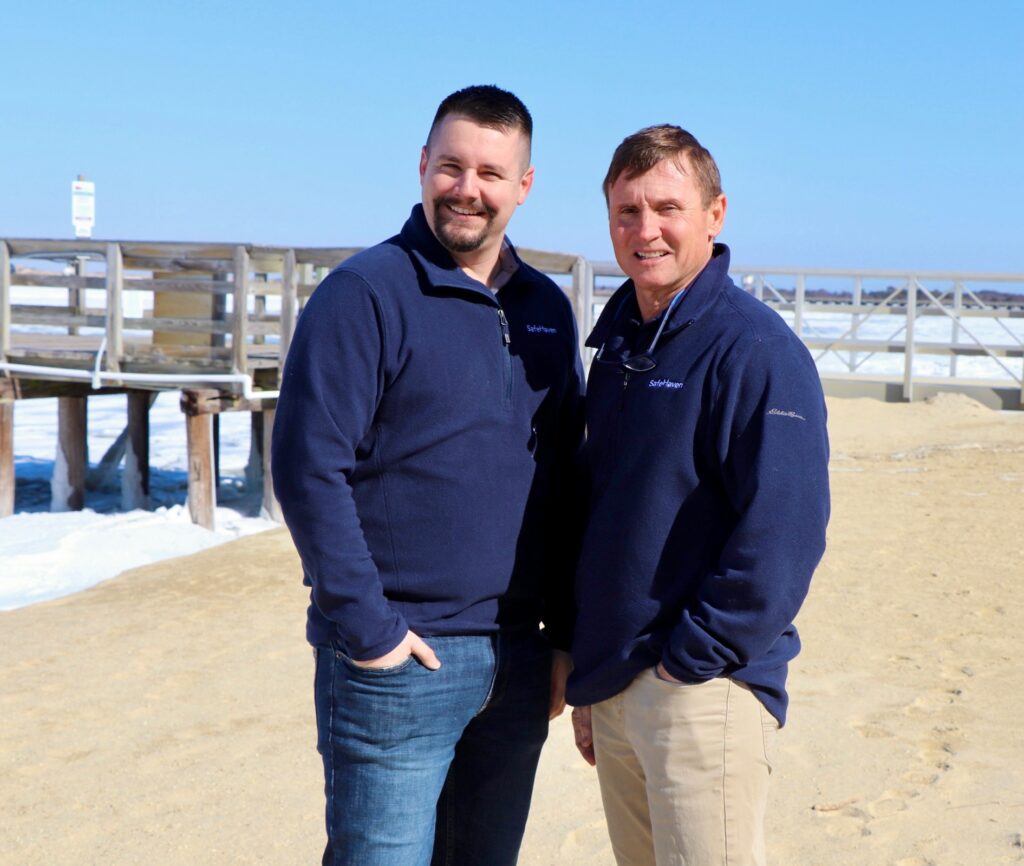 Two men wearing navy blue pullovers stand side by side on a sandy beach with a wooden pier and water in the background under a clear blue sky. Both are smiling and facing the camera.
