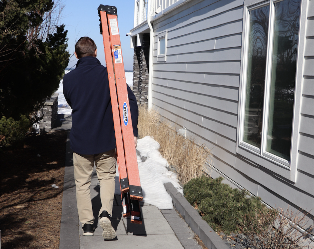 A person in a navy jacket and khaki pants carries an orange ladder along a narrow sidewalk beside a gray house, with patches of snow and shrubs lining the path on a sunny day.